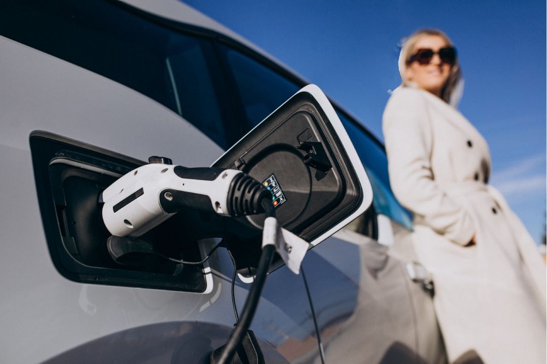 A hybrid car being charged with a charging cable connected to its port, with a woman standing in the background wearing a white coat under a blue sky.